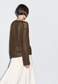 Bruine gebreide trui met lange mouwen en geribbelde zoom, gecombineerd met een vloeiende witte rok, gezien van achteren tegen een lichte achtergrond.