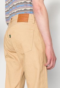 Pantalones de mezclilla beige con pierna recta y bolsillos traseros. Presentan un parche de cuero marrón con el logo en la cintura. Camisa a rayas gris y azul visible.