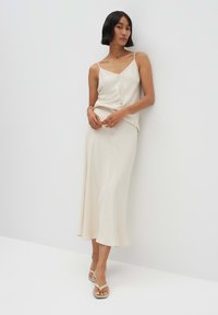 Light beige satin camisole with thin straps, paired with a matching ankle-length skirt. Model accessorized with a silver necklace, standing against a white wall.