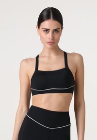 Reggiseno sportivo nero con spalline sottili e una fascia ampia. Presenta una bordatura bianca lungo il fondo e un tessuto liscio ed elastico.