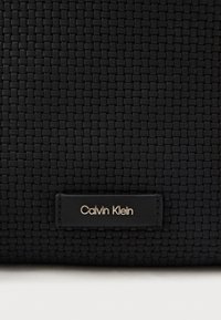 Μαύρη πλεκτή τσάντα χειρός με λεία υφή, διαθέτει ορθογώνια ετικέτα λογότυπου Calvin Klein σε ασημί υλικό πάνω σε δερμάτινη επιφάνεια.