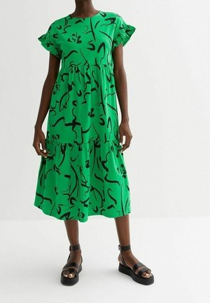 Kobieta w zielonej sukience midi z czarnymi abstrakcyjnymi wzorami i falbankowymi krótkimi rękawami, w połączeniu z czarnymi sandałami na platformie.