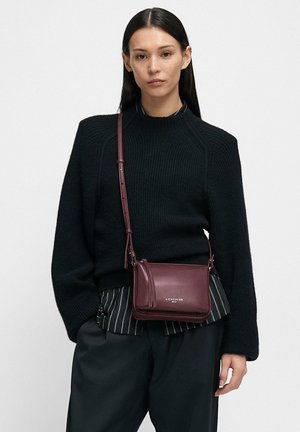 Woman wearing a black ribbed sweater, striped shirt, black pants, and carrying a small burgundy crossbody bag with a tassel detail.
