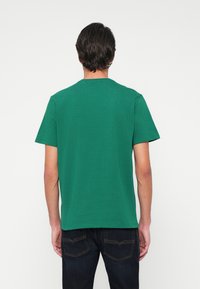 Vyras, vilkintis paprastą žalią trumpą rankovę marškinėlį ir tamsias džinsus, stovi veidu į baltą fono sieną, atsukęs nugarą žiūrovui.