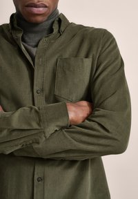 Olivgrünes Hemd mit Knöpfen, featuring eine Brusttasche, strukturierten Stoff und langen Ärmeln. Getragen über einem dunklen Rollkragenpullover.