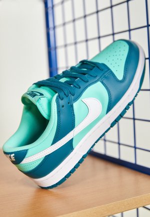 But sportowy Nike w odcieniach morskiego i niebieskozielonego z białym znakiem, umieszczony na drewnianej powierzchni na tle niebieskiej siatki.