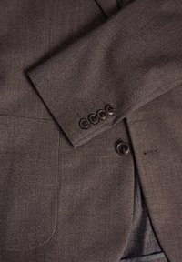 Smeđi tweed sako s teksturiranom površinom, s reverom, dva prednja džepa i pet smešnih dugmadi na manžetama.