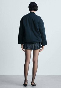 Giubbotto bomber nero, tessuto morbido, vestibilità rilassata; abbinato a pantaloni corti scuri con volant e collant neri trasparenti; design minimalista, senza decorazioni visibili.