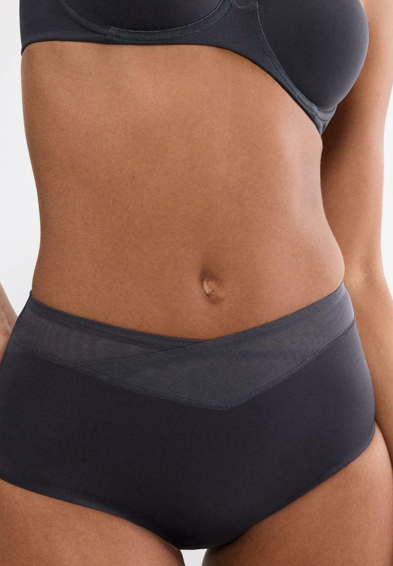 Vrouw draagt een zwarte high-waist onderbroek met meshdetails en een bijpassende zwarte beha, weergegeven van de taille tot de heupen tegen een witte achtergrond.