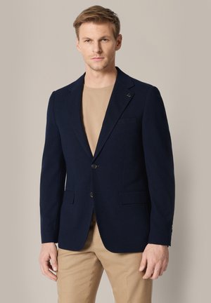 Mężczyzna o krótkich jasnobrązowych włosach, ubrany w granatową marynarkę na beżowej koszuli i beżowych spodniach, stojący na jednolitym, neutralnym tle.