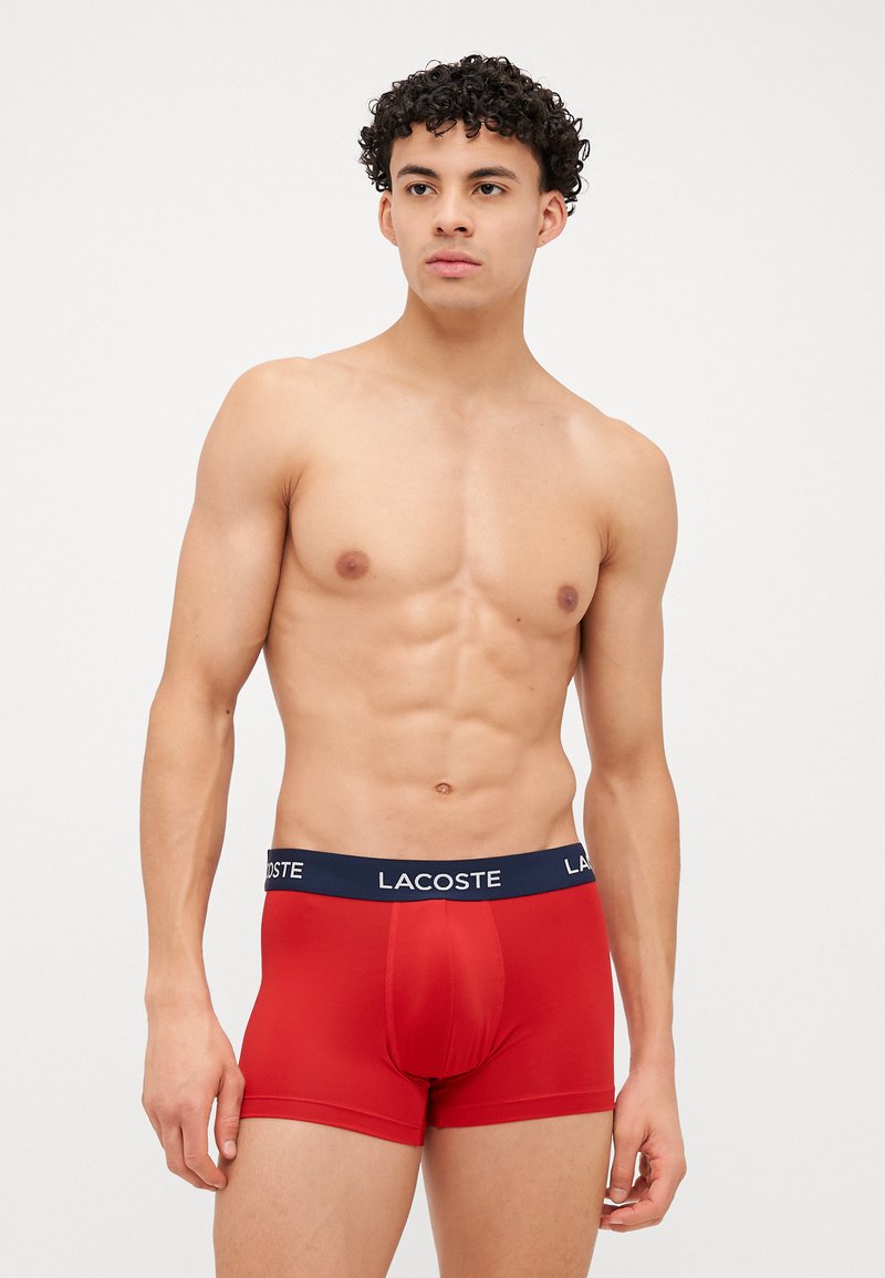Boxers vermelhos elásticos com uma cintura azul-marinho apresentando o logótipo "Lacoste" em branco. Superfície suave e design ajustado.