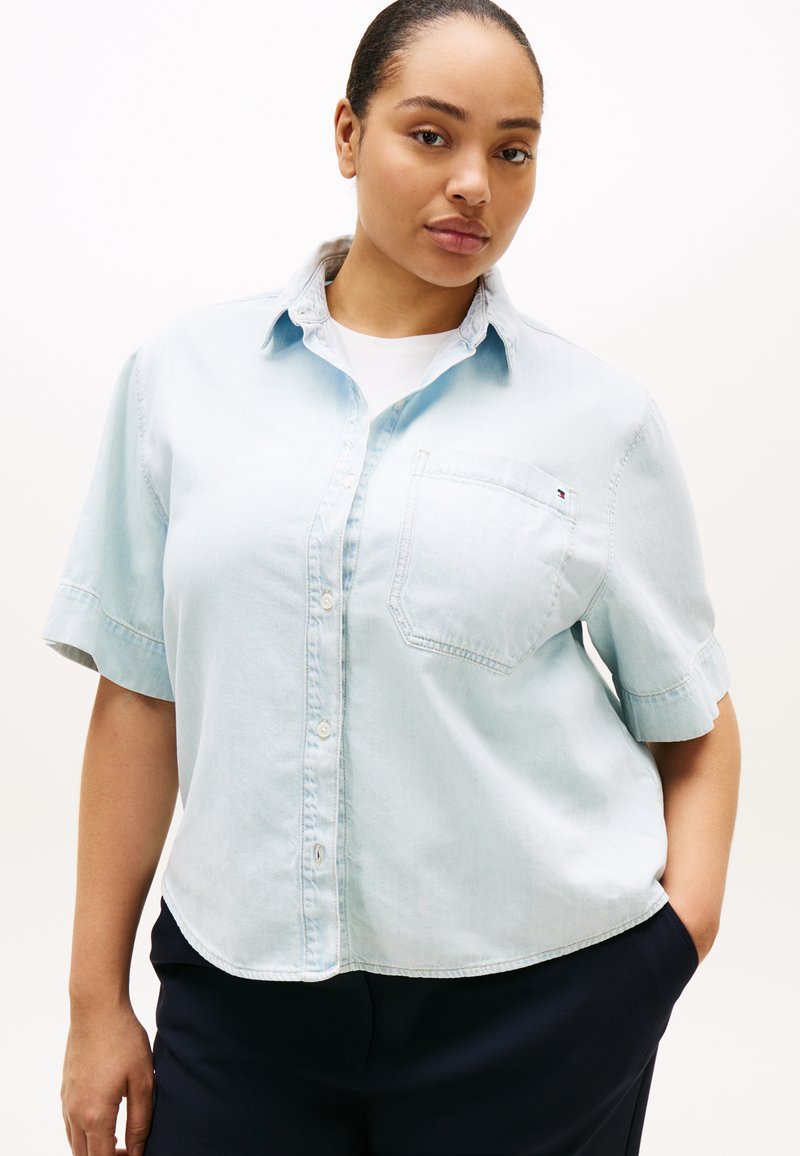 Γυναίκα που φοράει ένα ανοιχτό γαλάζιο πουκάμισο με κοντά μανίκια πάνω από ένα λευκό τοπ και σκούρα παντελόνια, πόζαροντας με το ένα χέρι στην τσέπη.