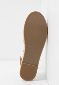 Semelle d'une chaussure beige avec un fond en caoutchouc texturé et une inscription de taille 4, inclinée contre un mur d'angle blanc.