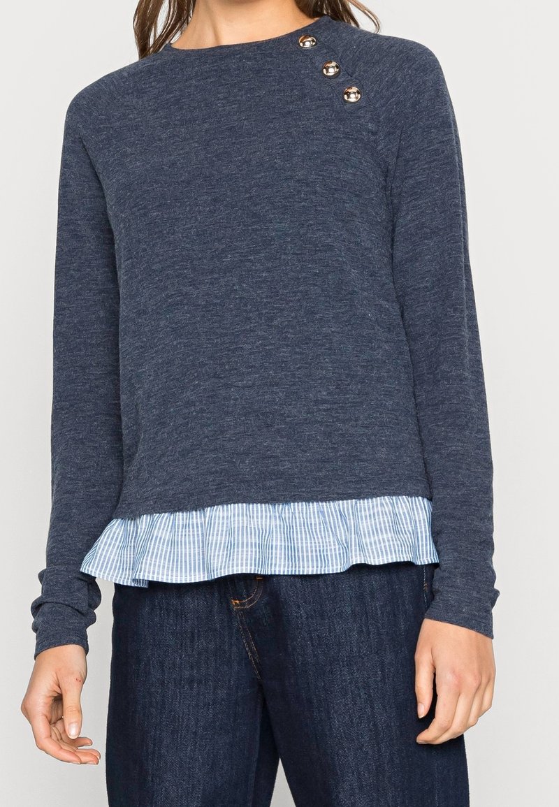 Femme portant un pull à manches longues bleu marine avec trois boutons métalliques sur l'épaule et un ourlet à volants rayé bleu et blanc.