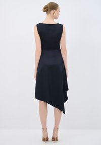 Robe en satin noir avec un design sans manches, ourlet asymétrique, texture lisse et forme minimaliste, associée à des talons nus.