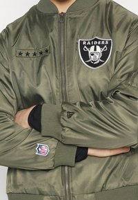 Olive green bomber jacket with ribbed cuffs, featuring a Raiders logo and stars patch on the chest, satin-like material, zip front.
