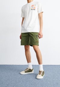 T-shirt crème avec une poche graphique colorée, associée à un short cargo vert avec des zippers, et des baskets olive avec des accents blancs.