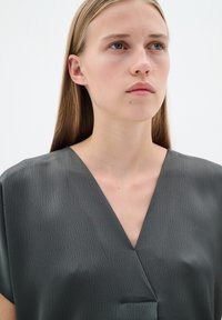 Bluză gri texturată cu decolteu în V, mâneci scurte și o croială lejeră. Material neted cu un model geometric, având o strălucire subtilă.