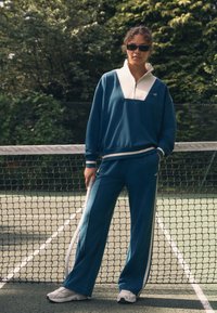 Tuta da ginnastica in pile turchese con mezza cerniera, colletto bianco e strisce laterali bianche, abbinata a scarpe da ginnastica bianche su un campo da tennis.