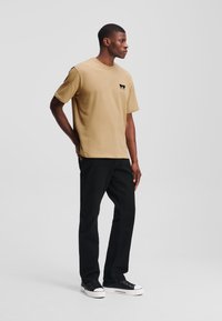 Beige lühikese varrukaga T-särk ümarate kaelustega ja väikese musta logoga, koos mustade sirgete pükste ja mustade tossudega.