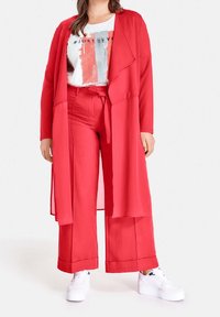 Mujer vistiendo un abrigo largo rojo y pantalones anchos a juego, con una camiseta gráfica blanca y zapatillas de plataforma blancas.