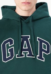 Green cotton hoodie featuring large "GAP" embroidery in navy with white outline. Includes a front pocket and drawstring hood.