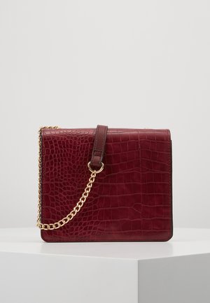 Bordeauxrote Handtasche aus künstlichem Krokodilleder mit goldener Kettenriemen, die auf einem weißen Podest vor einfachem Hintergrund liegt.