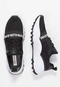 Ett par svarta och vita träningsskor med Calvin Klein Jeans-logotypen överst och tjocka, strukturerade sulor på en vit bakgrund.