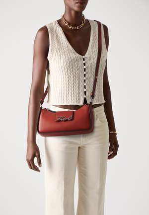 Red leather shoulder bag with silver hardware, featuring a silver logo and contrasting edges. Worn with a knit tank and light trousers.