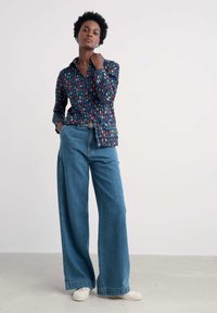 Koszulka w niebieskim, czerwonym i białym wzorze, zestawiona z jasnoniebieskimi spodniami typu wide-leg. Posiada kołnierzyk i zapięcie na guziki. Całość dopełniają białe trampki.