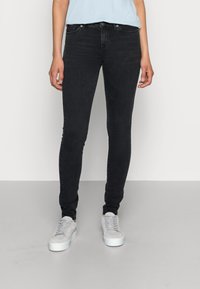 Jeans skinny pretos em denim, com cintura média, design de cinco bolsos e detalhes costurados, combinados com ténis cinzentos claros.