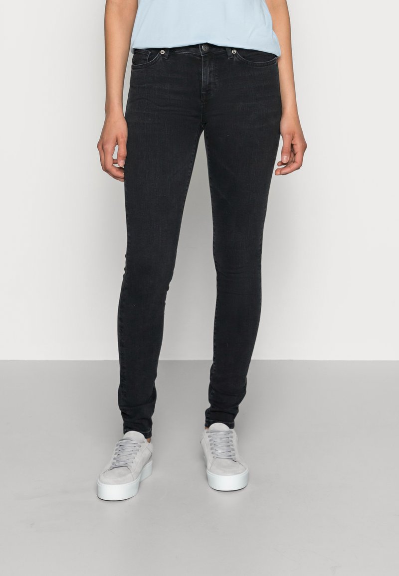 Jeans skinny pretos em denim, com cintura média, design de cinco bolsos e detalhes costurados, combinados com ténis cinzentos claros.