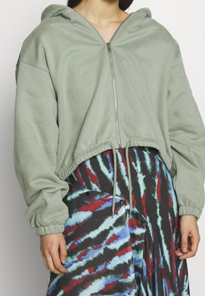 Sweat à capuche court en tissu vert discret avec une fermeture éclair, un ourlet resserré et des manches longues. Assorti à une jupe à motifs multicolores.