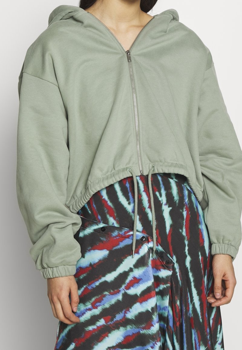 Sudadera capucha de corte ajustado en tela verde apagado con cremallera, dobladillo fruncido y mangas largas. Combinada con una falda de patrón multicolor.