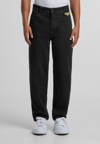 Pantalons en denim noir avec une coupe droite, design à cinq poches et accent logo jaune. Portés avec un débardeur blanc et des baskets blanches.
