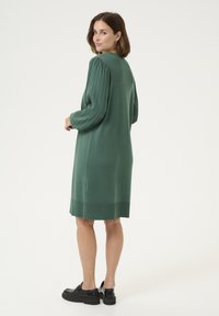 Robe tricotée verte avec de longues manches côtelées et une coupe droite, se terminant au-dessus du genou. Modèle associé à des chaussures noires épaisses.