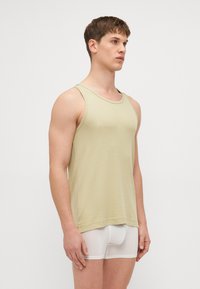 Débardeur sans manches vert clair avec une texture côtelée, décolleté rond et ourlet piqué. Porté par-dessus un boxer blanc.