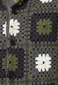 Cardigan croșetat cu un design din patchwork, având flori gri, verzi și albe, nasturi negri și detalii din fire texturate.