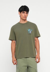 Oliven grøn bomulds T-shirt med korte ærmer. Forsiden har et grafisk print af fugle og logo. Rund halsudskæring og afslappet pasform.