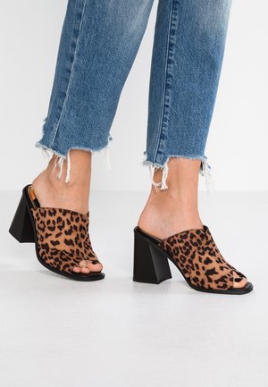 Mules de punta abierta con estampado de leopardo y tacón cuadrado. Parte superior de tela suave, suela negra y lados abiertos, combinados con jeans azules desgastados.