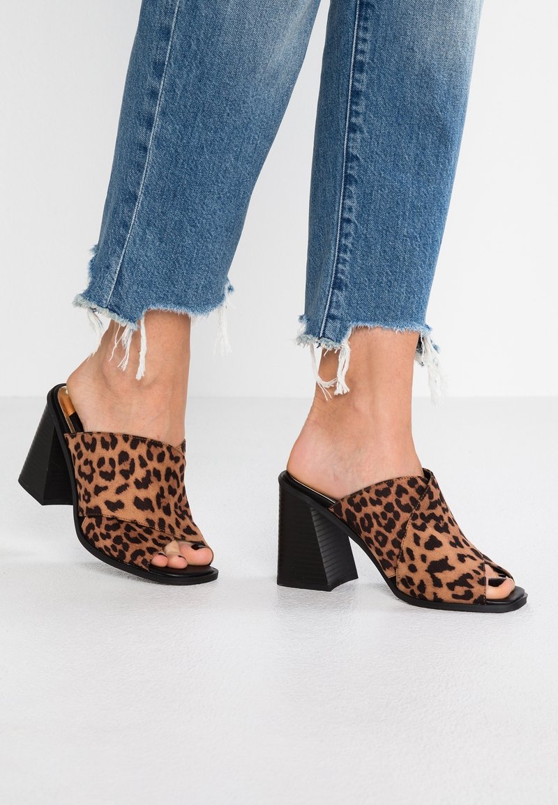 Mules à bout ouvert en imprimé léopard avec un talon bloc. Dessus en tissu doux, semelle noire et côtés ouverts, associés à un jean bleu délavé.