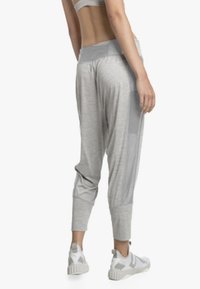 Vrouw in lichtgrijze joggerpants, witte sport-bh en witte sportschoenen, staand met haar rug naar de camera tegen een witte achtergrond.