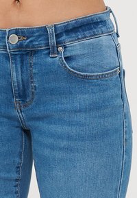 Blå denimjeans med en tettsittende design, som har en toppknapp, glidelåsåpning og frontlommer. Stoffet har en glatt tekstur med lett falming.
