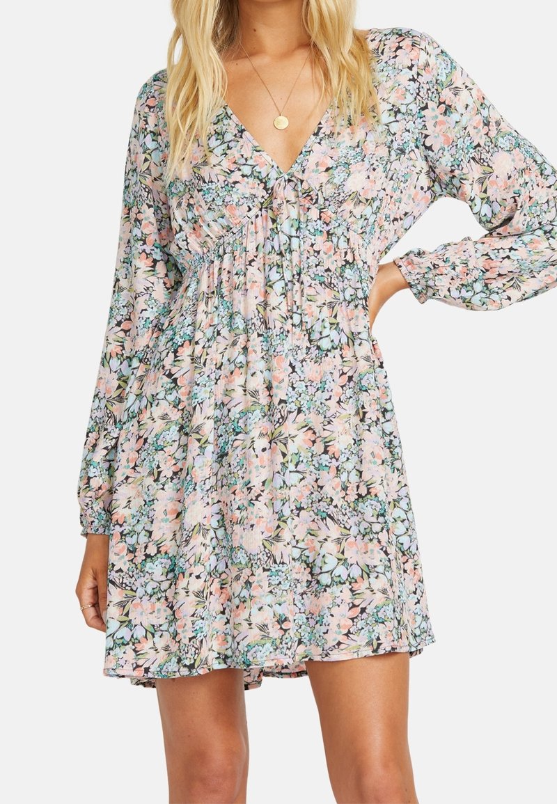 Femme portant une robe longue à manches longues florale pastel avec un décolleté en V et une taille froncée, accessoirisée d'un collier pendentif rond en or.