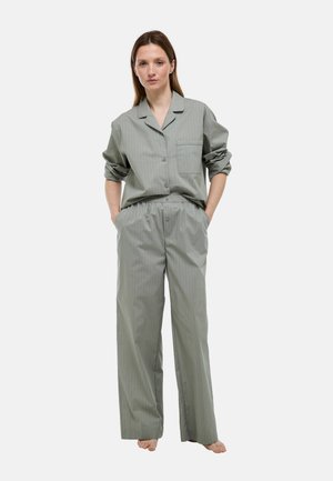 Femme debout pieds nus portant une chemise ample à fines rayures grises claires boutonnée et un pantalon large assorti, les mains dans les poches.