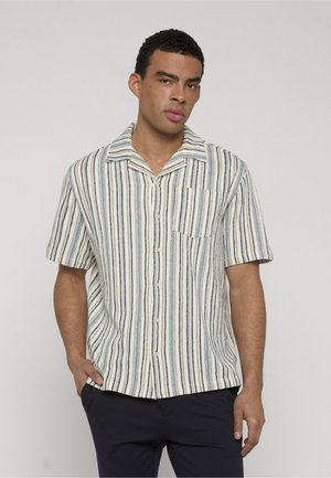 Mężczyzna w koszuli na krótki rękaw z pionowymi, niebieskimi, czarnymi i beżowymi paskami, stojący z jedną ręką w kieszeni na jednolitym tle.