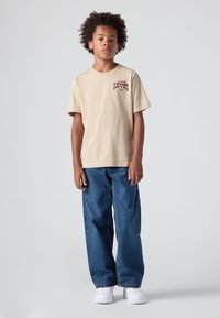 Bež majica iz bombaža z maroon logotipom. Nošena z ohlapnimi modrimi kavbojkami in belimi teniskami. Preprost dizajn, nevtralne barve.