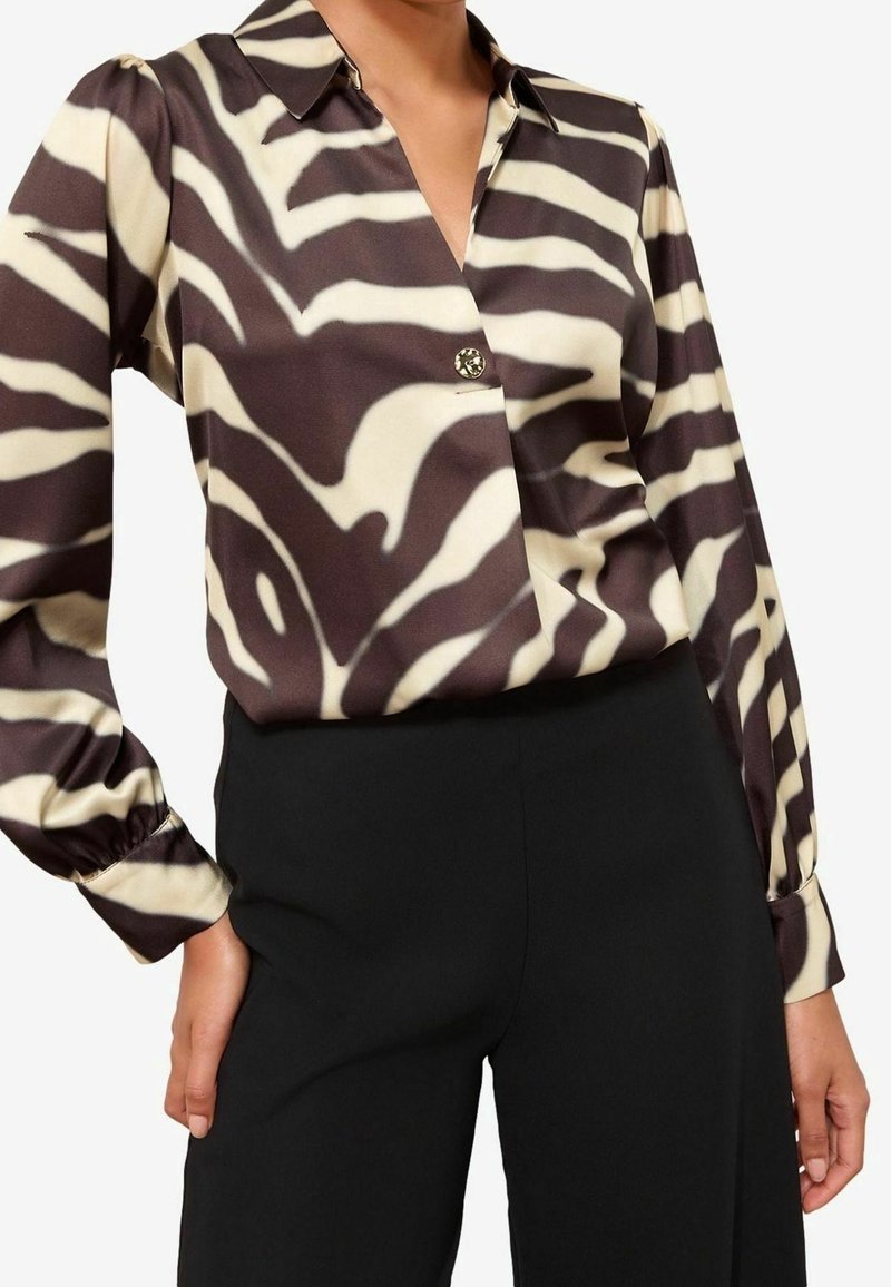 Nainen pukeutuneena mustavalkoiseen seepra-kuvioiseen pitkähihnaiseen puseroon, joka on laitettu korkeavyötäröisten mustien housujen sisään.