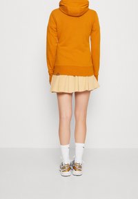 Felpa arancione con cappuccio abbinata a una gonna pieghettata beige, calze bianche e sneakers grigie con dettagli dorati. L'outfit è casual e sportivo.