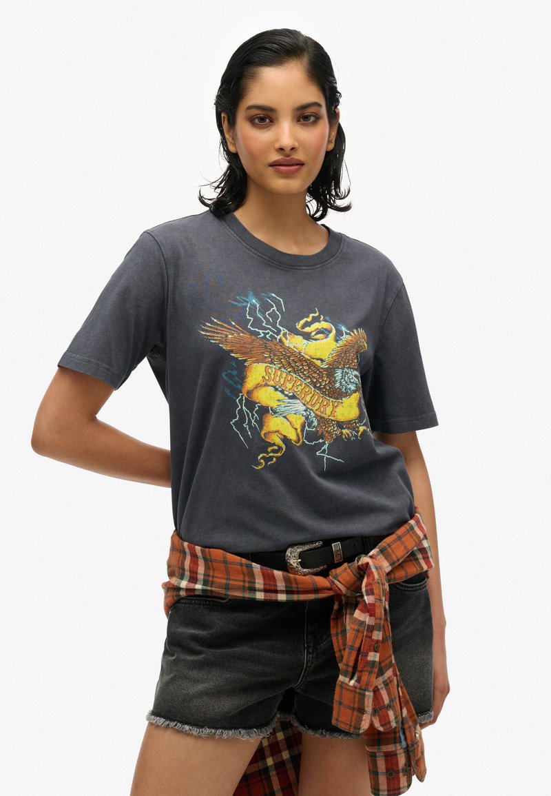 Camiseta gris de manga corta con un gráfico colorido de un águila y texto. Combinada con shorts de mezclilla oscuros y una camisa de cuadros naranja atada a la cintura.